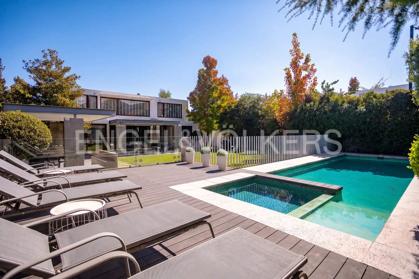 A modern house with a pool and lounge chairs on a sunny day. The pool is turquoise and surrounded by a wooden deck.