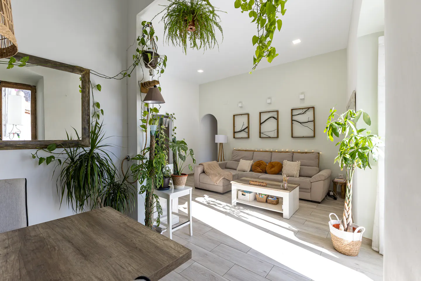 Bright living room with light wood floors, beige sofa, and many plants. A wooden table is in the foreground.