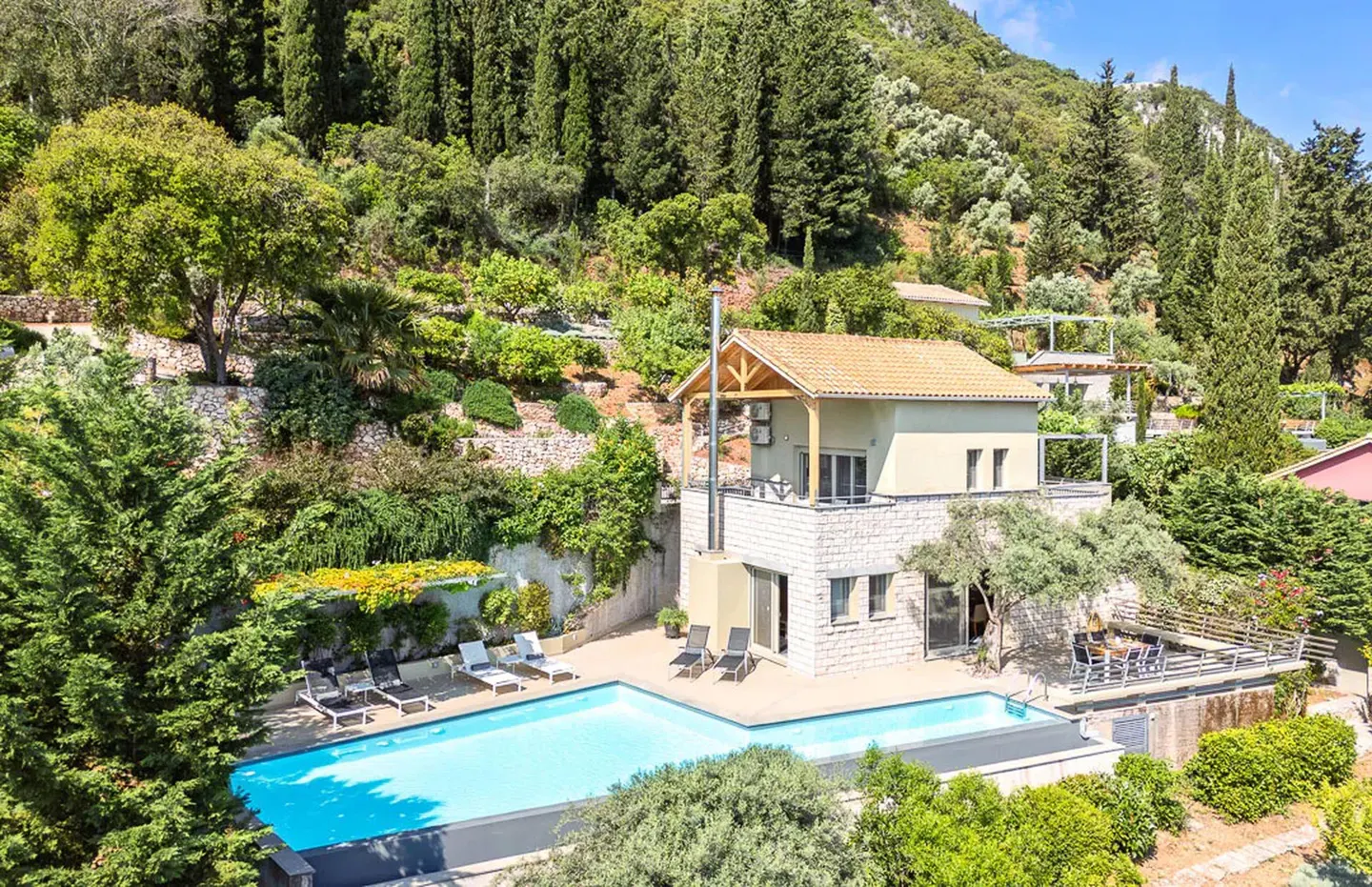 Two-story stone villa with a private pool, sun loungers, and a lush, terraced hillside backdrop.