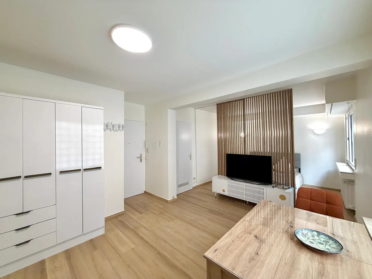 Modern studio apartment with light wood floors, white wardrobe, TV on a white console, and a wooden slat room divider.