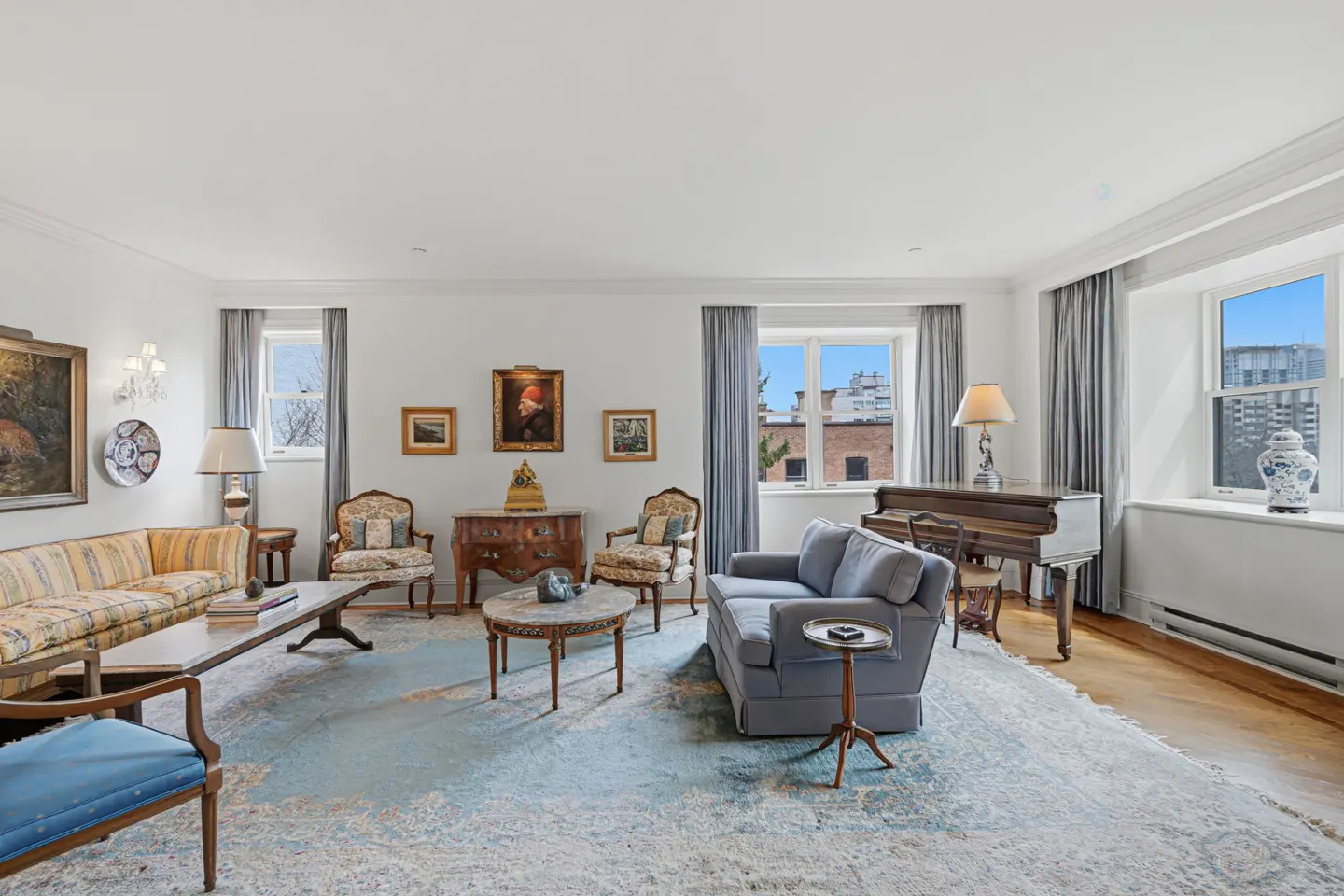 Bright living room with a blue rug, a gray sofa, a piano, and antique furniture. Windows with gray curtains let in natural light.