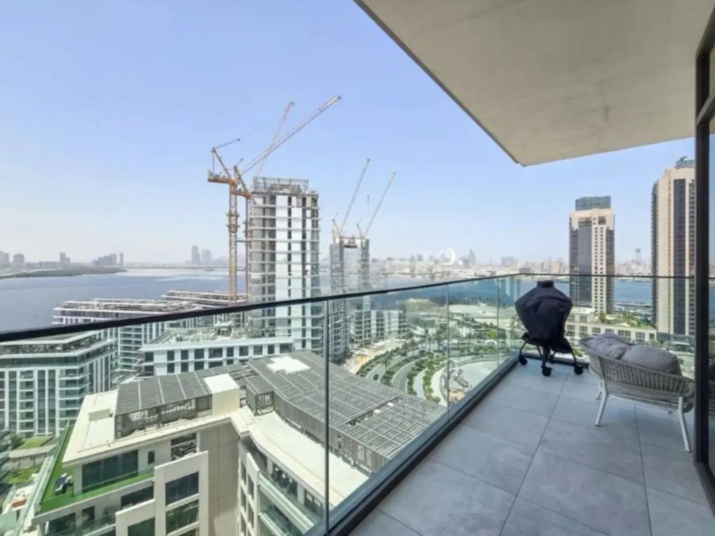 Balcony view of a city skyline with cranes, buildings, and water. A grill and chair sit on the tiled balcony.
