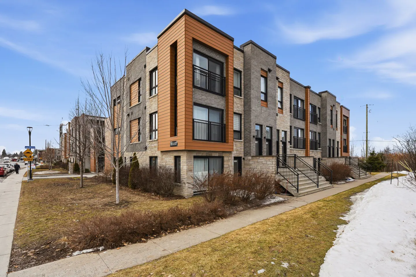 Exterior view of modern townhouses with gray brick, brown accents, black railings, and concrete steps on a partly sunny day.