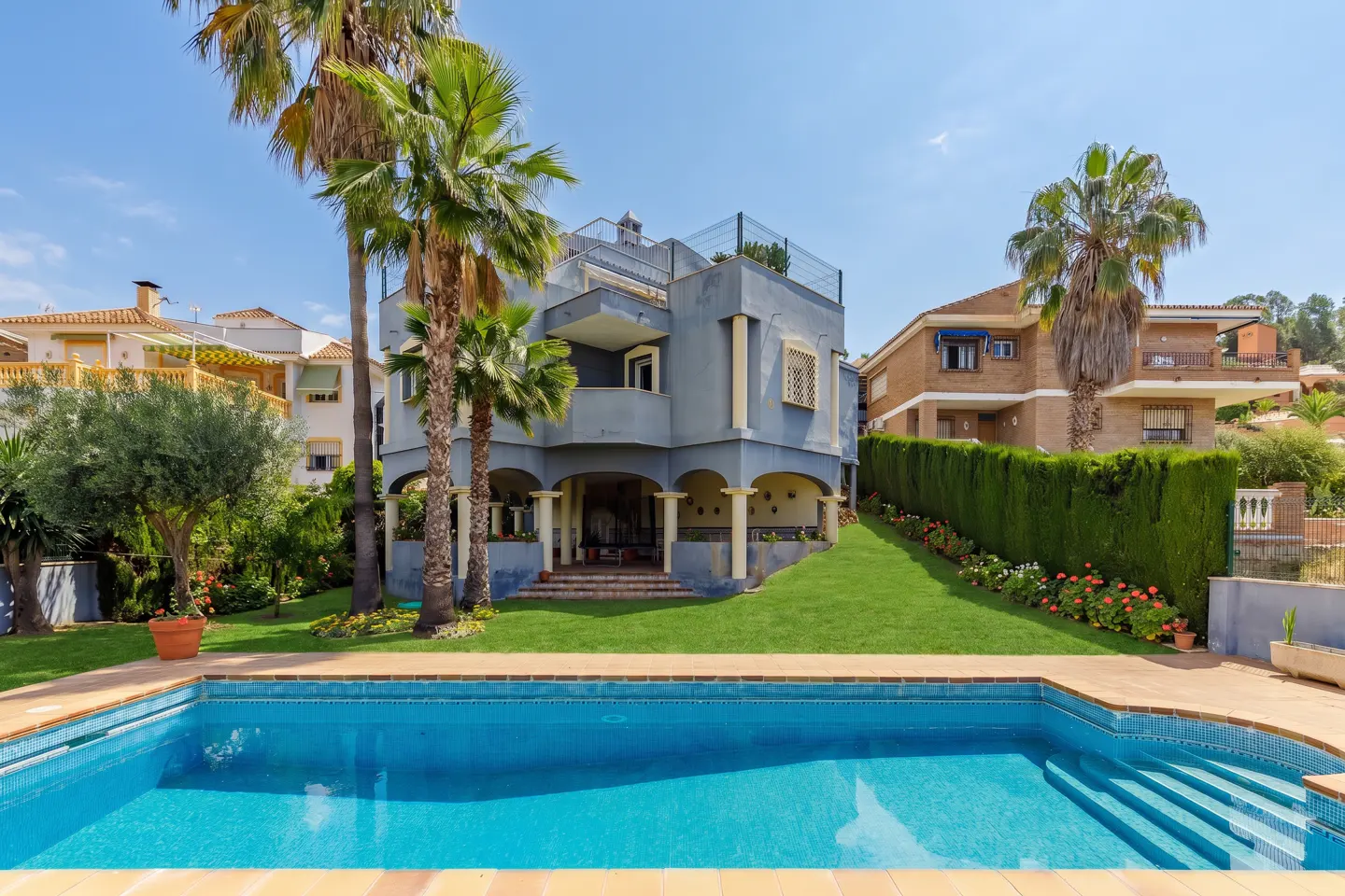 A blue swimming pool in the foreground, with a large blue house, palm trees, and green lawn in the background.