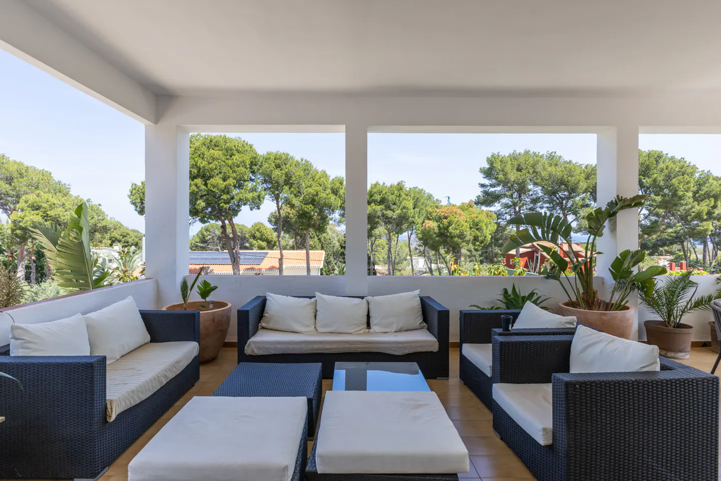 Outdoor patio with black wicker furniture, white cushions, and potted plants. Trees are visible in the background.