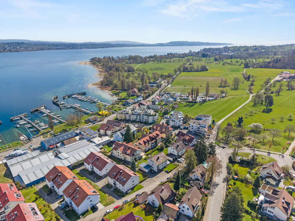 Aerial view of a lakeside town with red-roofed houses, a marina filled with boats, and green fields under a blue sky.