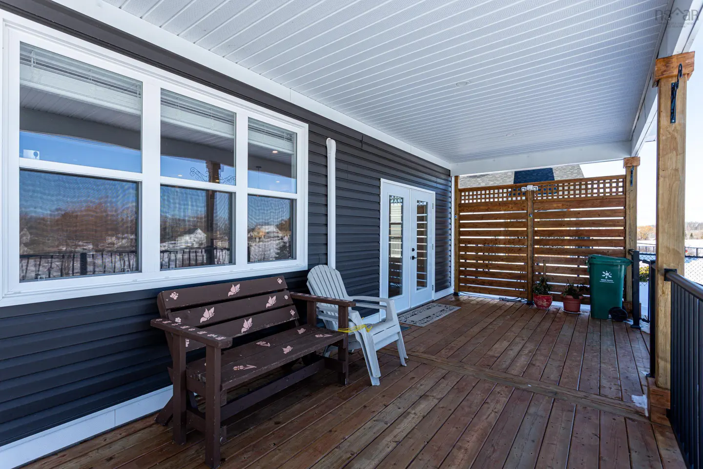 Covered wooden deck with a brown bench, white chair, and white french doors. A wooden lattice fence and green trash can are on the right.