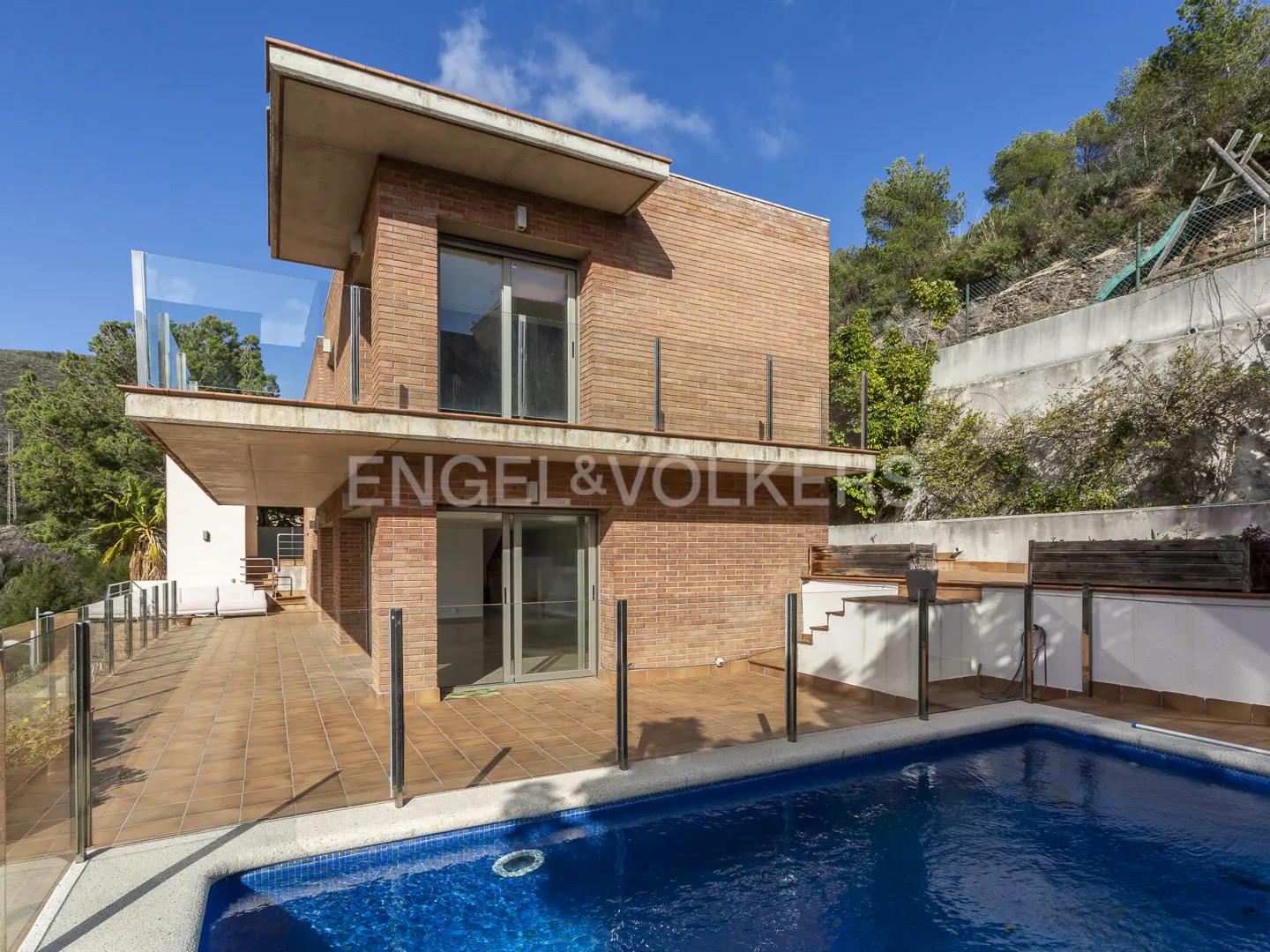 Modern two-story red brick house with a tiled patio, glass railings, and a blue swimming pool under a clear blue sky.