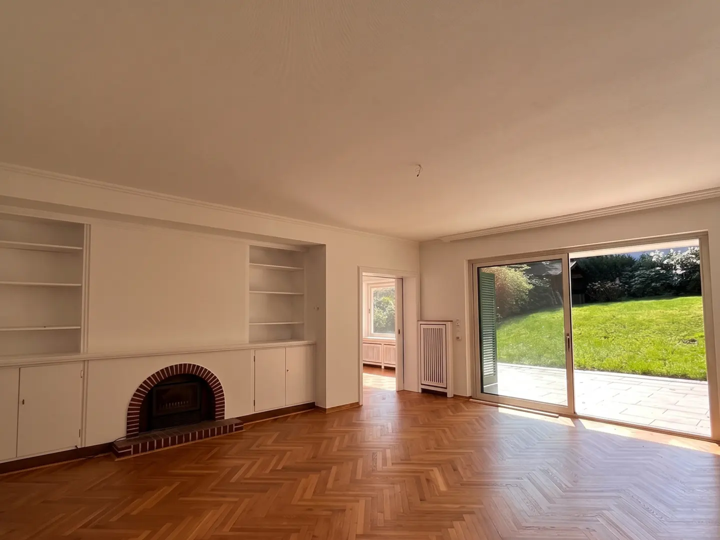 Bright, empty living room with herringbone wood floors, white walls, built-in shelves, fireplace, and sliding glass doors to a green lawn.
