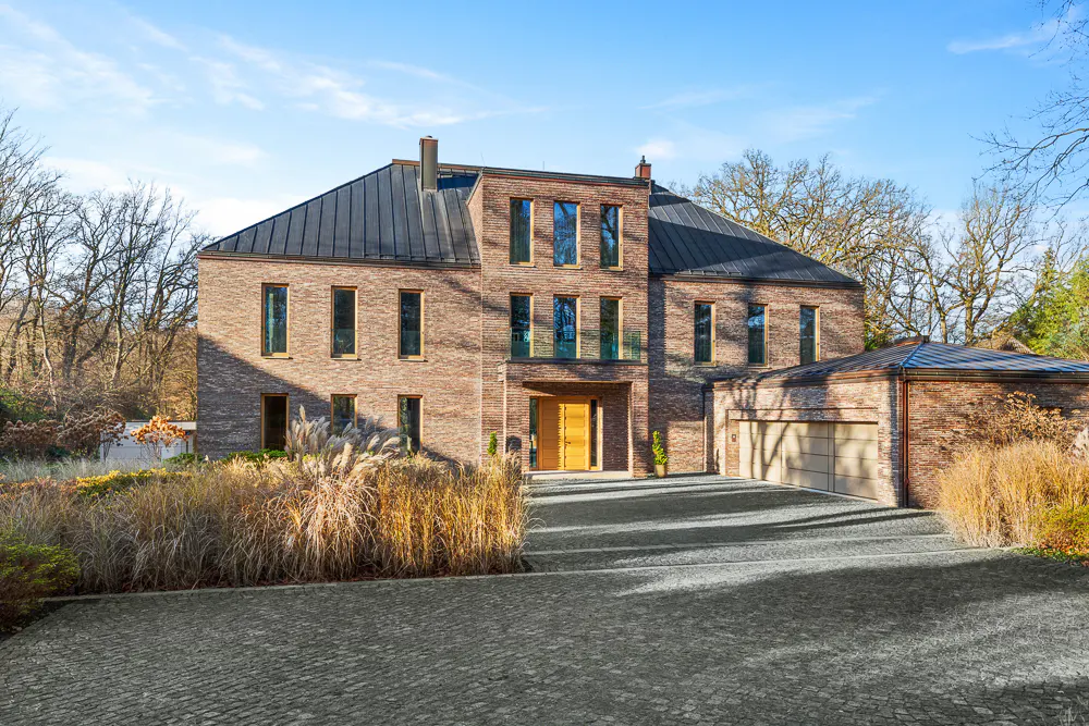 A three-story brick house with a black roof and a gray cobblestone driveway. Tall grass lines the driveway.