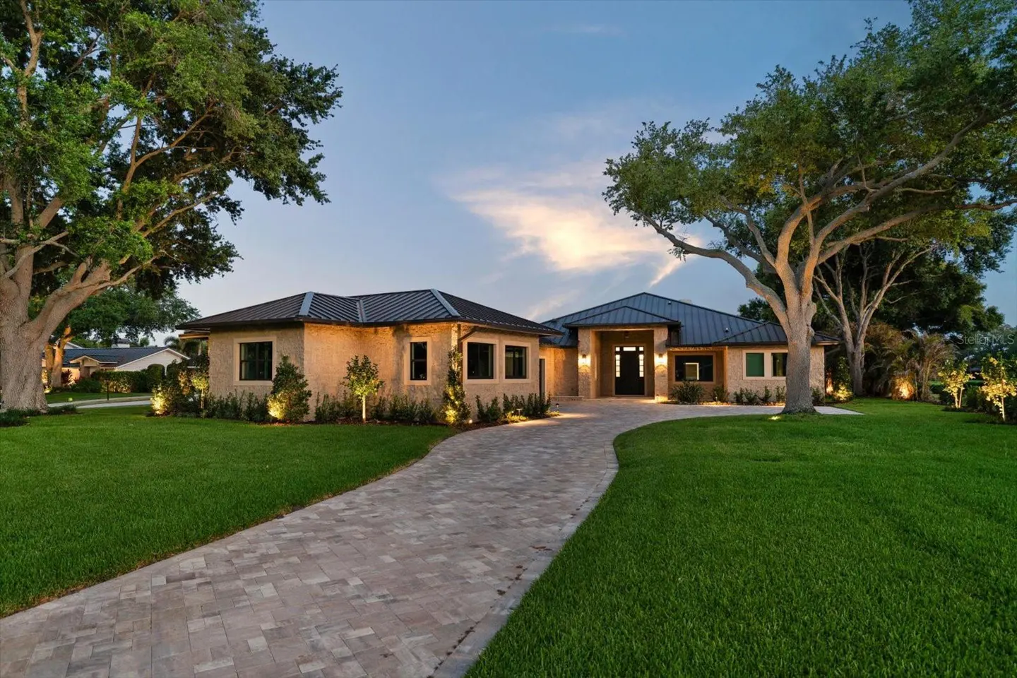 A tan, single-story home with a dark roof and a brick driveway, surrounded by green grass and trees.