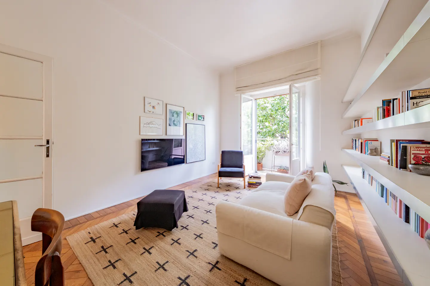 Bright living room with white sofa, black armchair, patterned rug, wall-mounted TV, and built-in bookshelves on parquet floor.