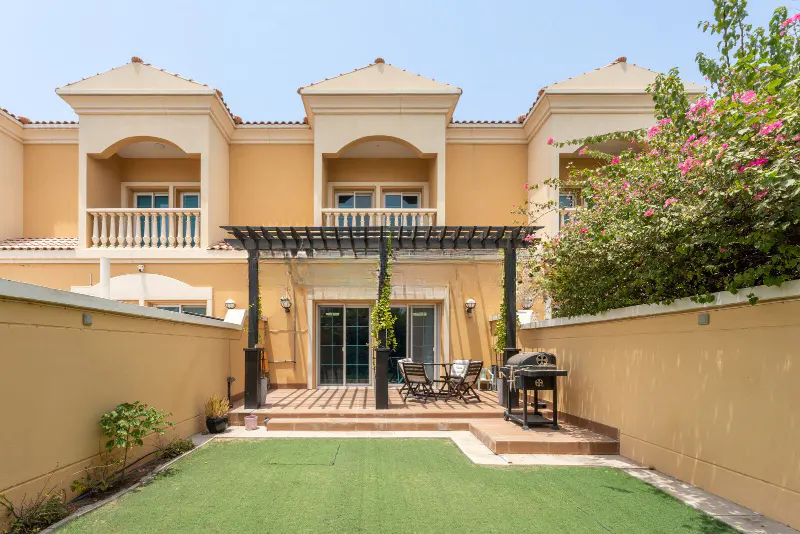 Backyard view of a tan two-story house with a patio, grill, table, chairs, and green lawn.