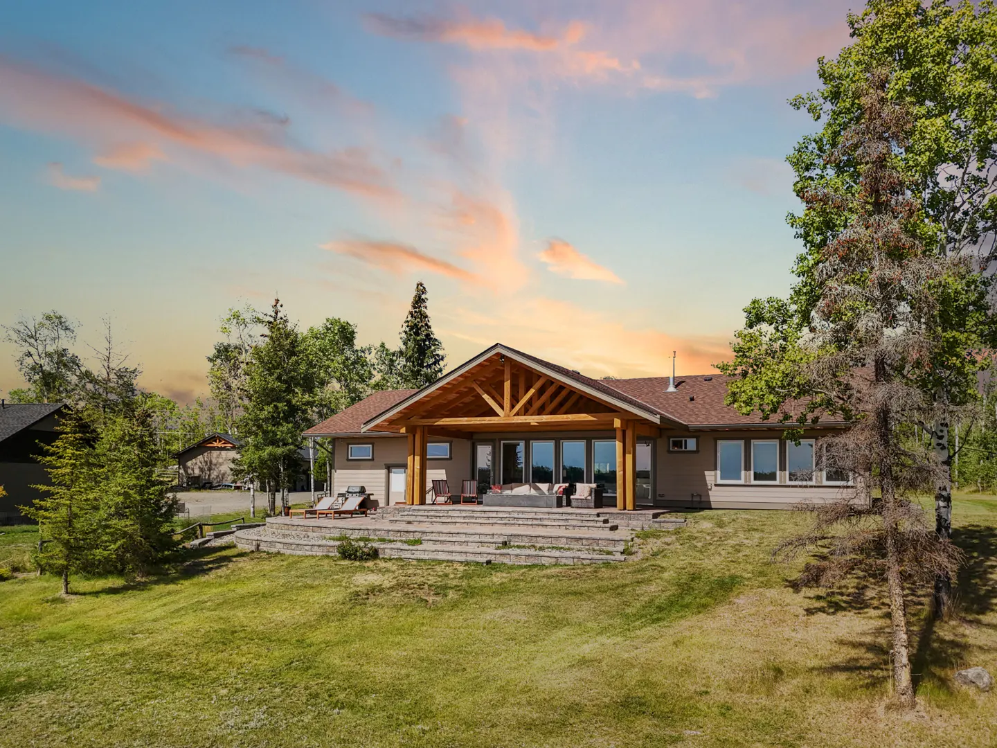 Exterior view of a beige house with a brown roof, a wooden patio cover, and a green lawn at sunset.