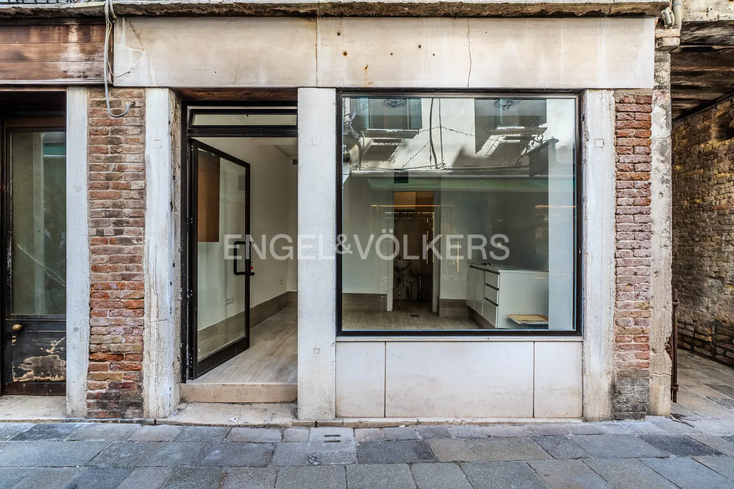 Exterior of a commercial space with an open glass door and large window. Engel & Völkers logo is on the window. The building is brick and stone.