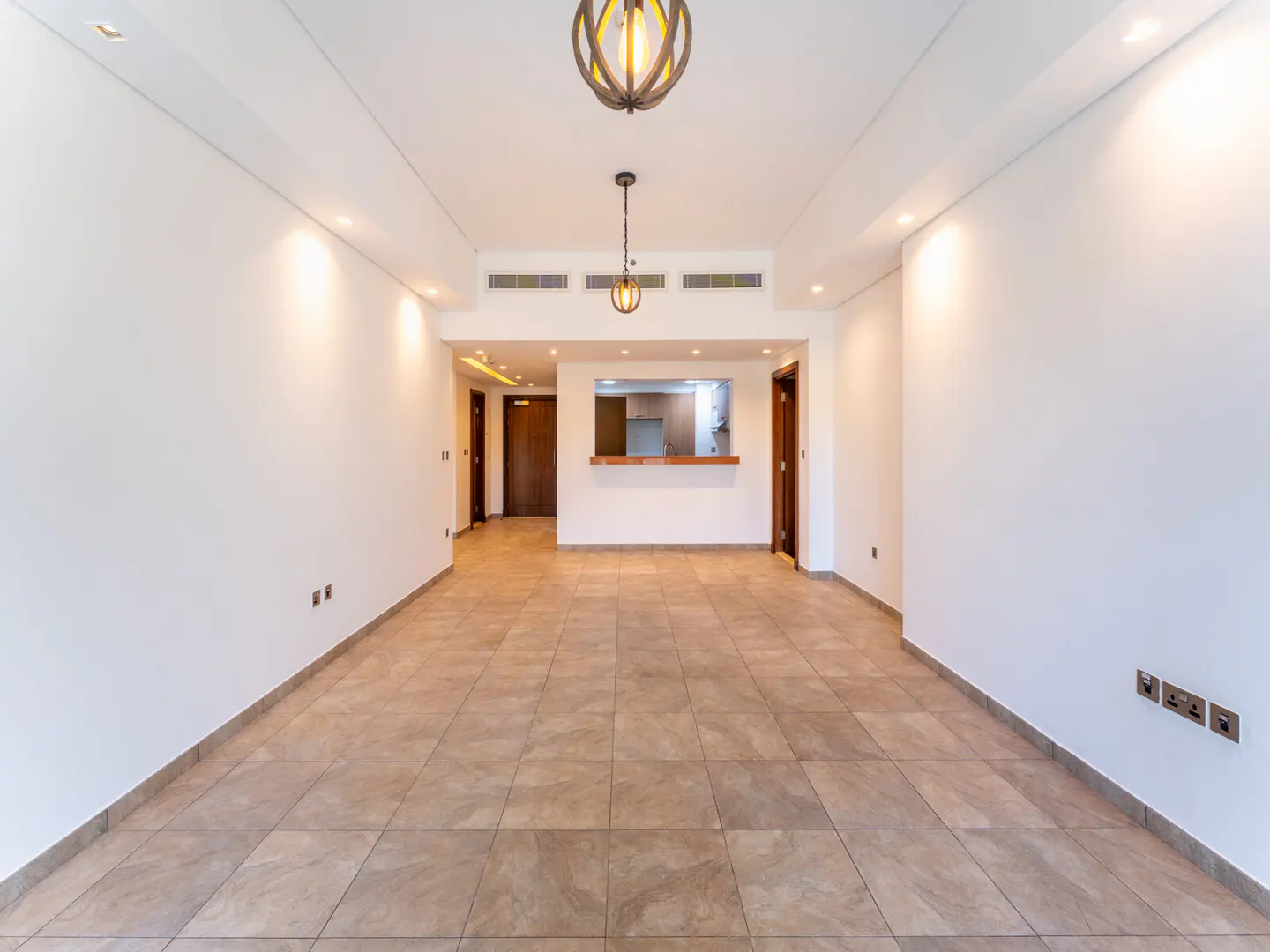 An empty room with white walls, tile floors, and two light fixtures. A kitchen is visible through a serving hatch.