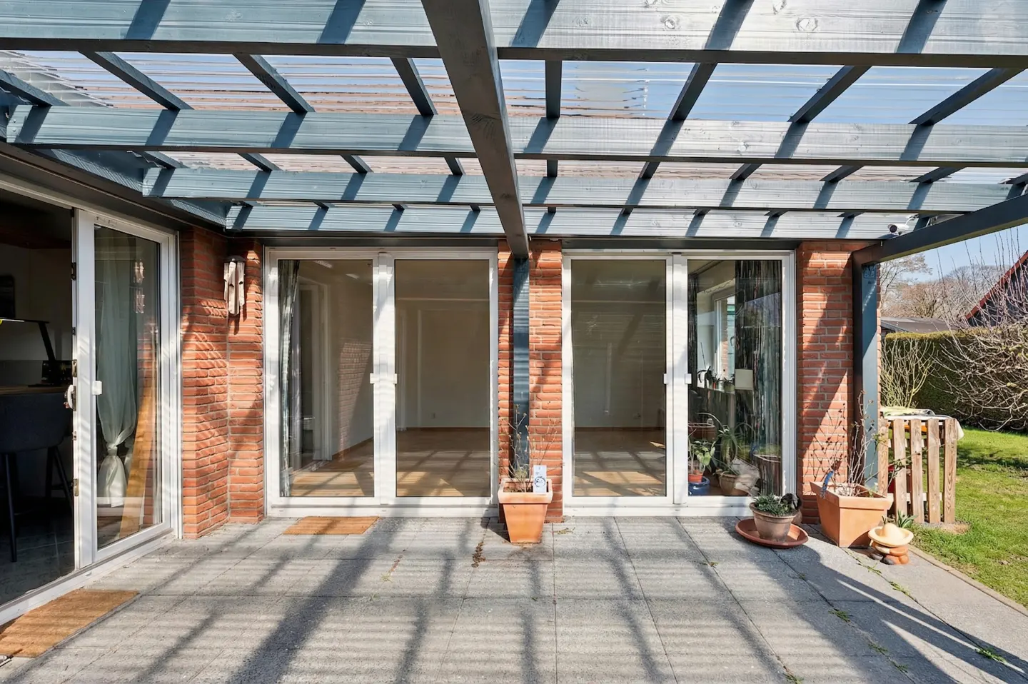 Patio with brick wall, sliding glass doors, and a pergola casting shadows on the gray stone floor. Potted plants add a touch of greenery.