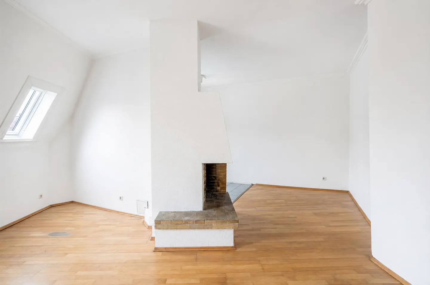 Bright, empty room with wood floors, white walls, and a brick fireplace. A skylight is visible on the left.