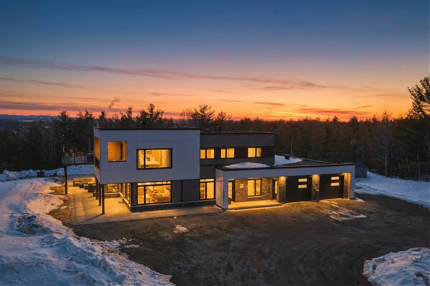 Modern two-story home with large windows, set against a sunset sky and snowy landscape.