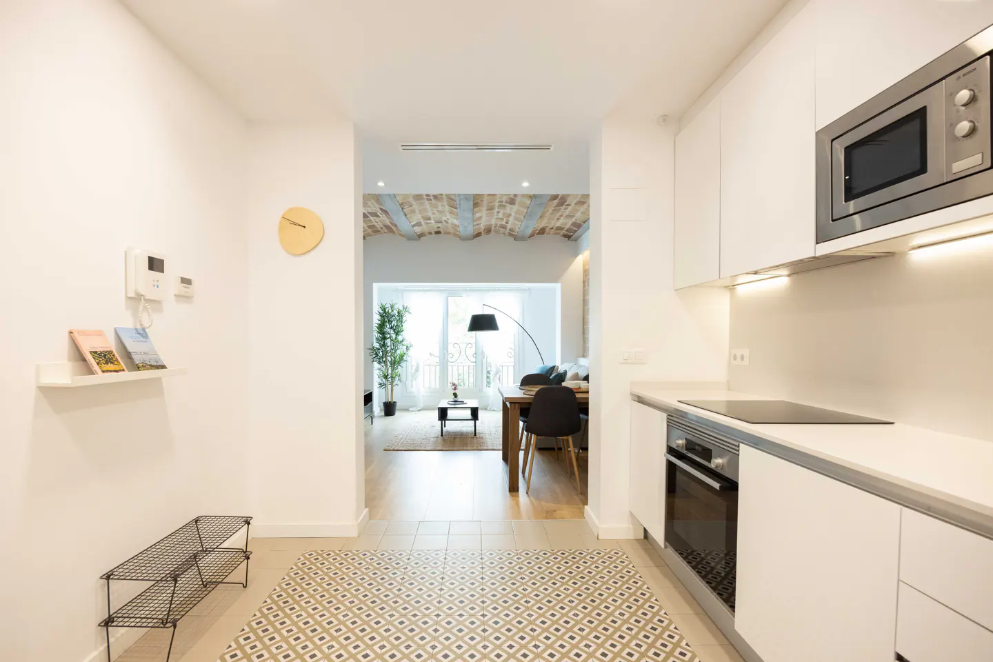 A bright, modern apartment interior with white walls, cabinets, and stainless steel appliances. A living room is visible through an archway.