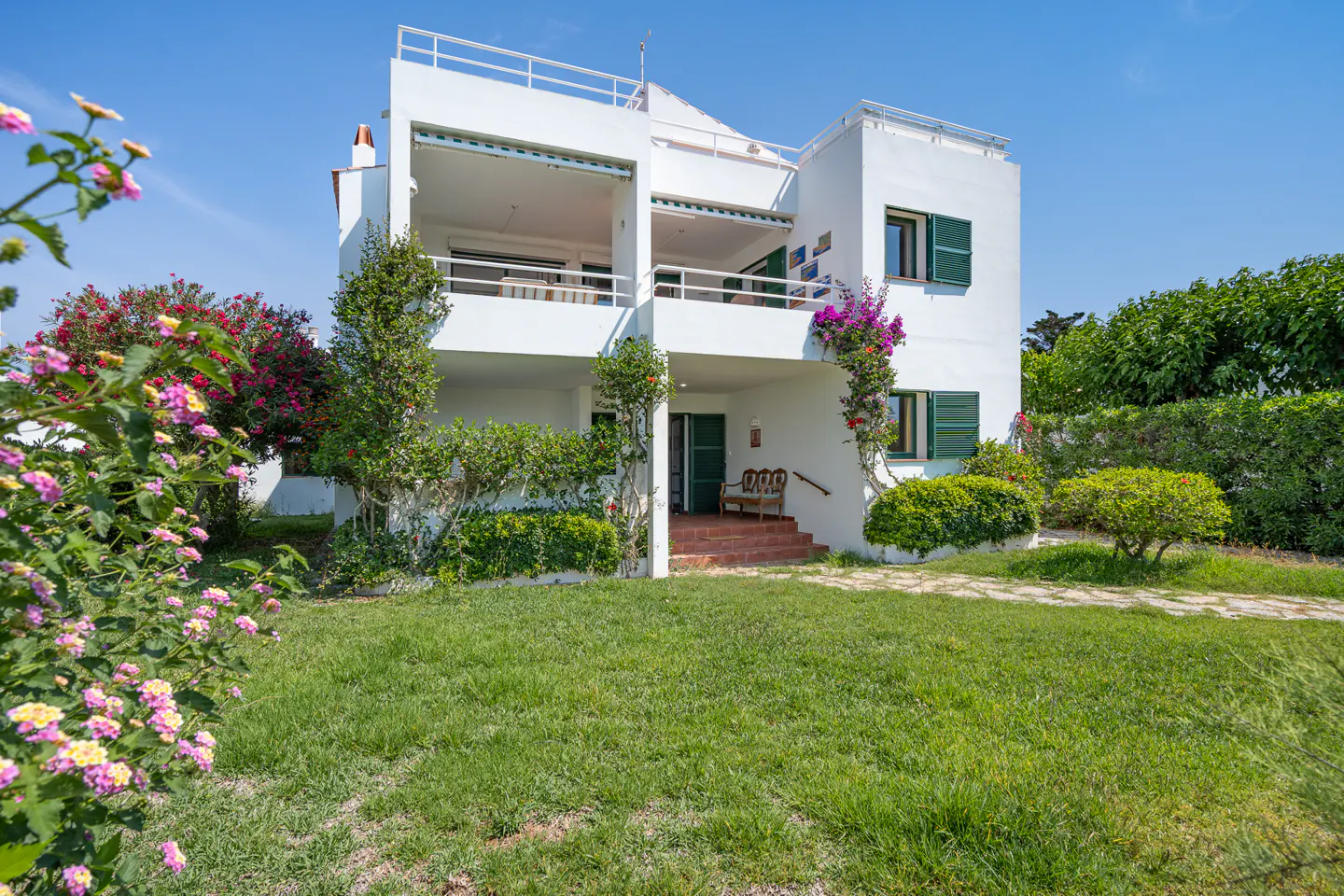 A white two-story Mediterranean villa with green shutters, a rooftop terrace, and a lush garden under a clear blue sky.