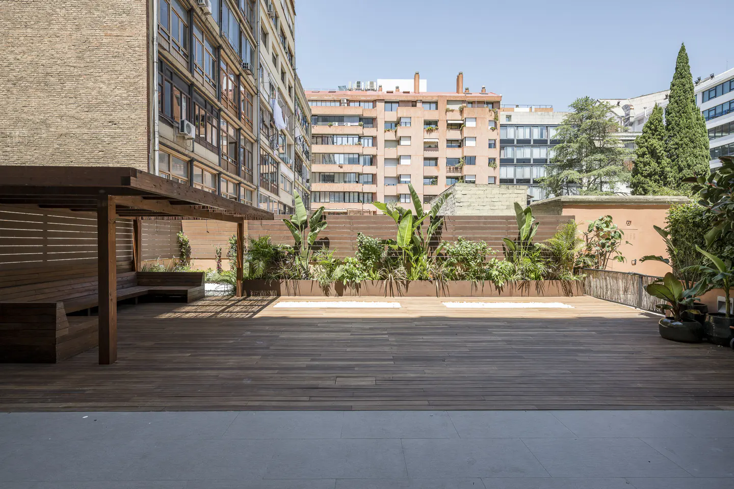Spacious rooftop terrace with wooden decking, a pergola, built-in seating, and lush green plants against city buildings.