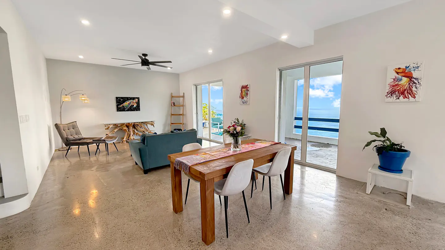 Bright living and dining area with ocean view. Table with flowers, blue sofa, and art on white walls. Balcony overlooks the sea.