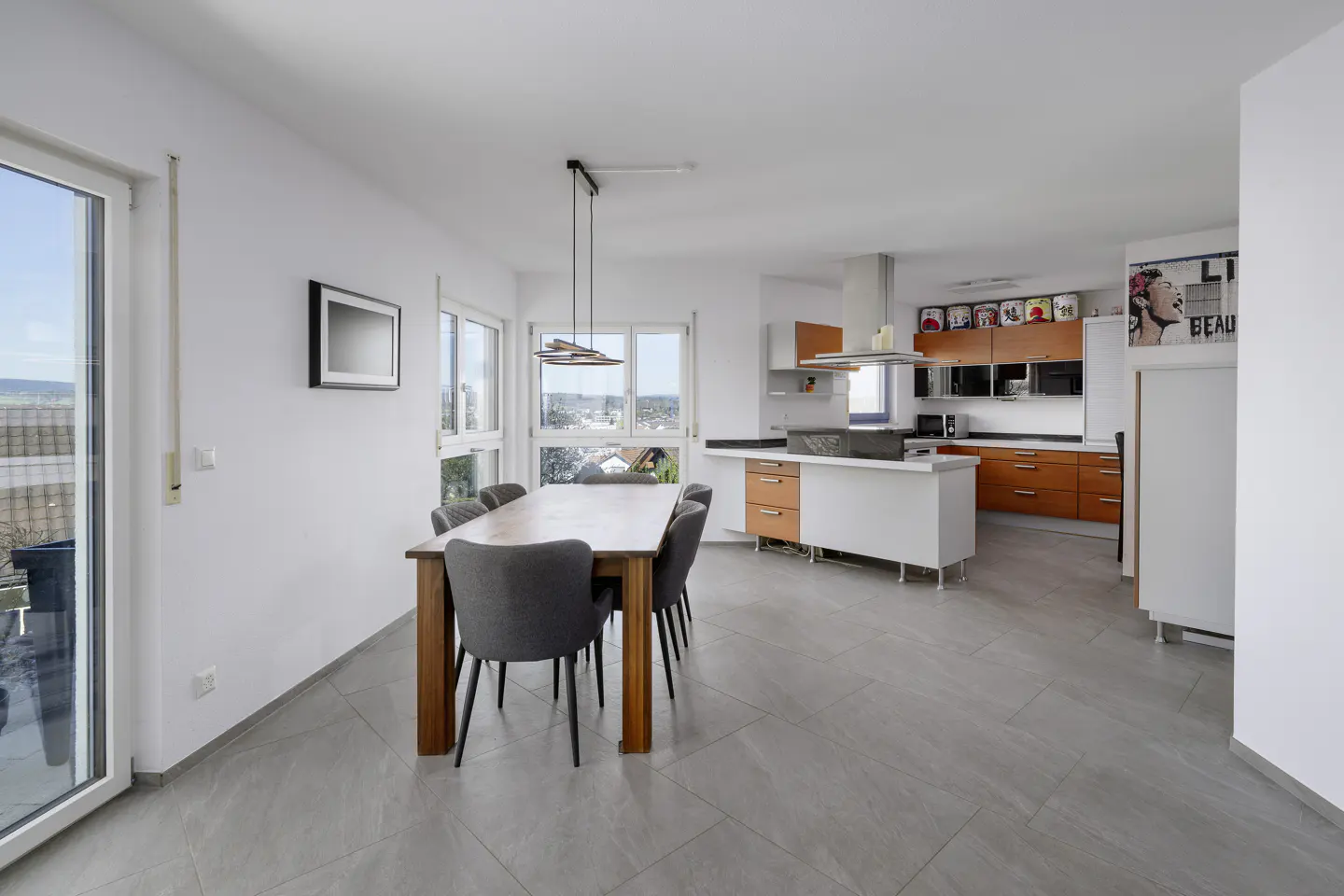 Open-concept kitchen and dining area with gray tile flooring. A wooden table with gray chairs sits near windows. Orange and white cabinets line the kitchen.