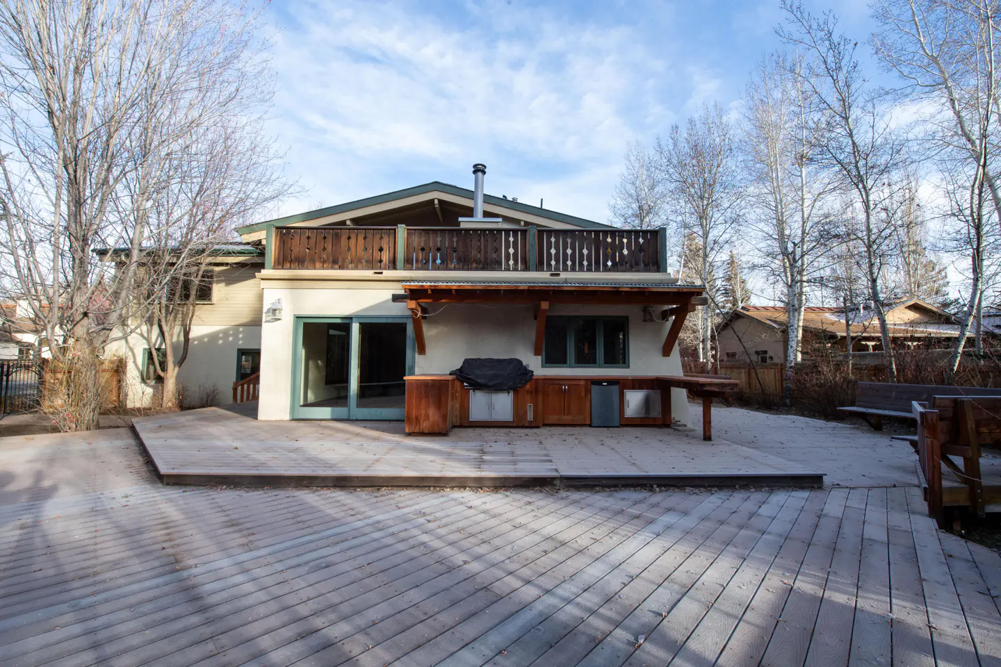Backyard view of a two-story house with a wooden deck, outdoor kitchen, and balcony. Bare trees surround the property.