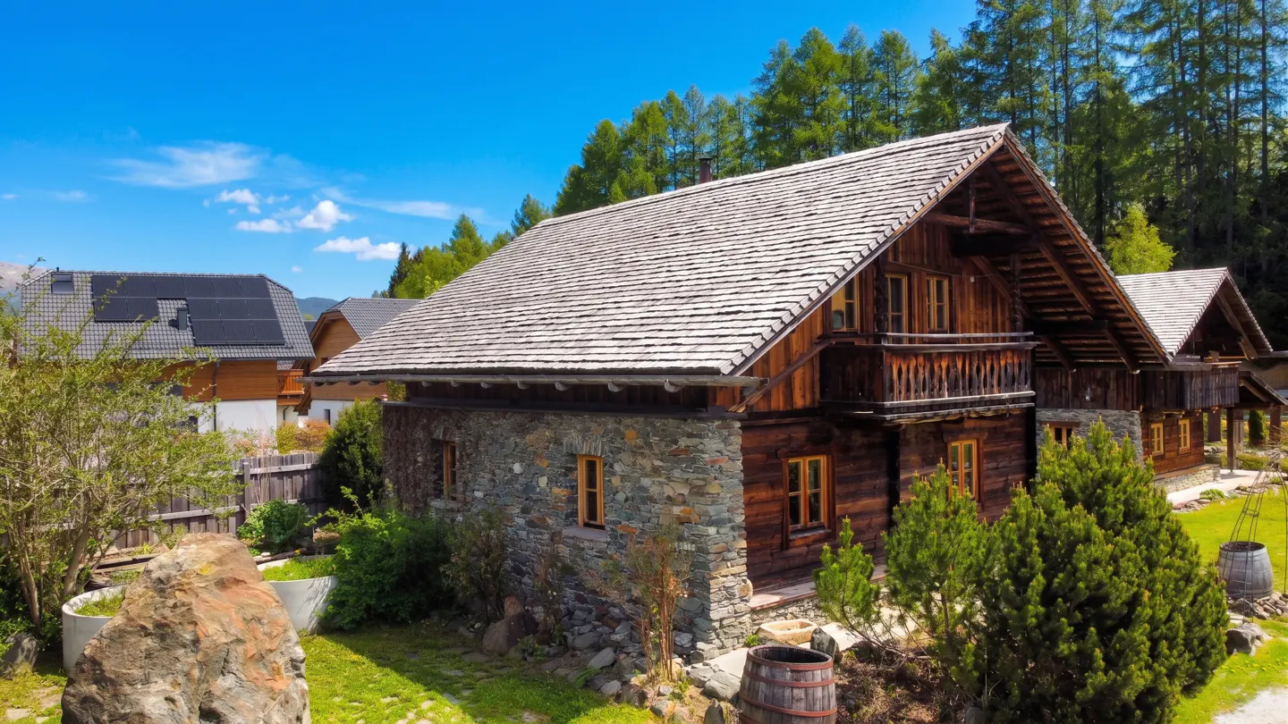 Rustic cabin with stone and wood exterior, gray shingle roof, and small balcony, set against a backdrop of green trees and blue sky.
