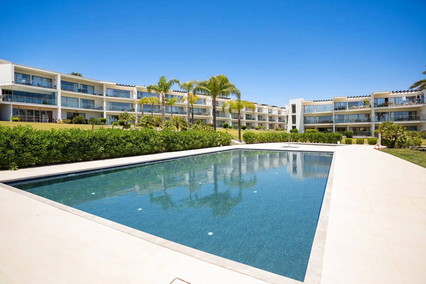A rectangular pool with blue water sits in front of white apartment buildings and palm trees under a clear blue sky.