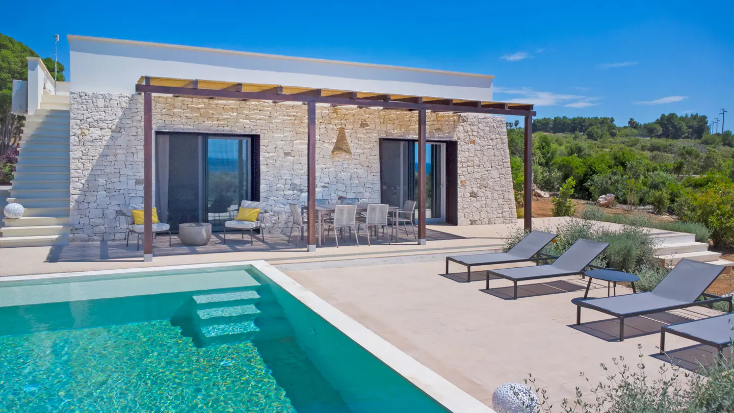 Modern stone villa with a swimming pool, patio lounge chairs, and a covered dining area under a clear blue sky.