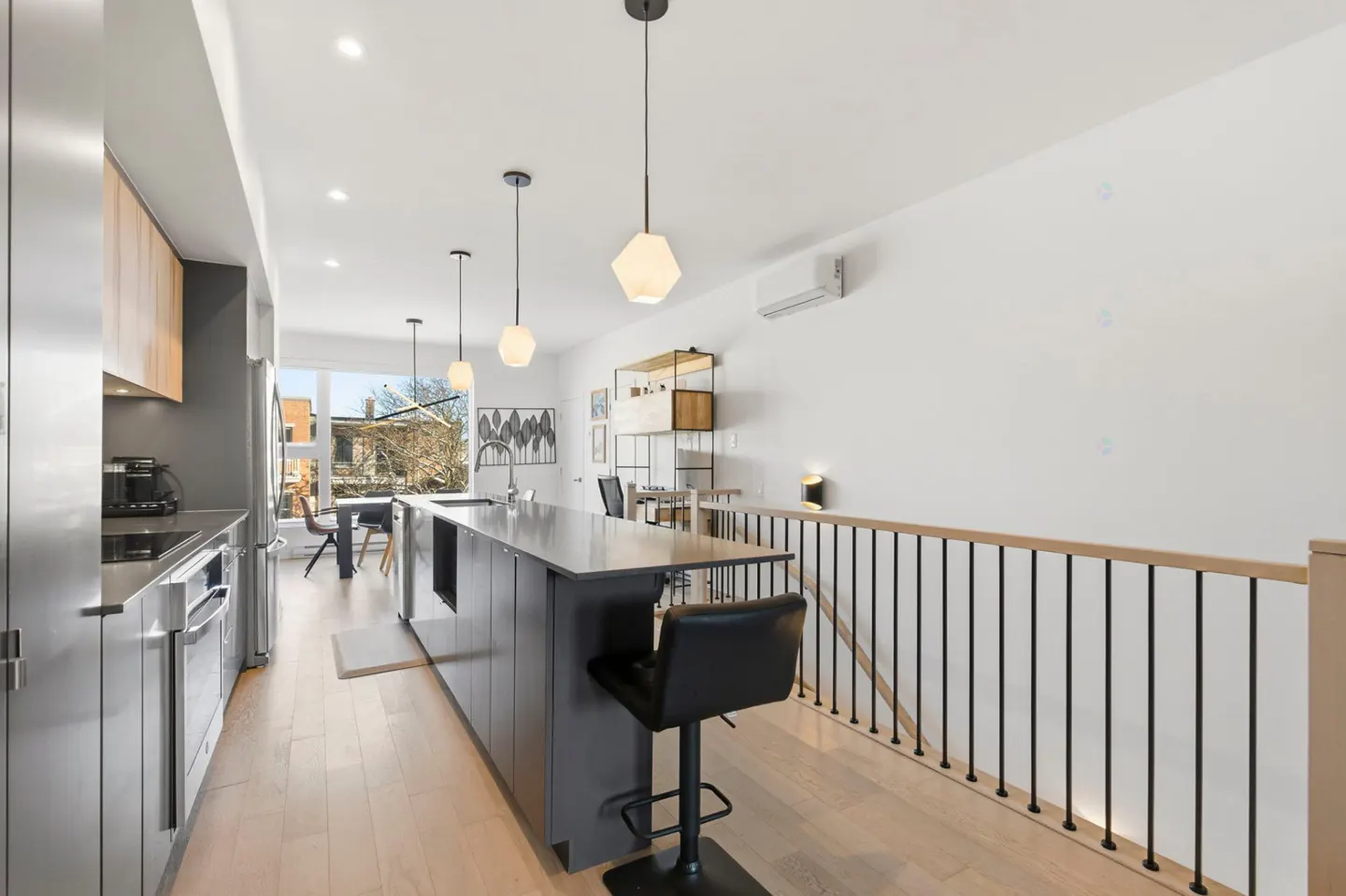 Modern kitchen with gray cabinets, stainless steel appliances, and a large island with a black stool. Hardwood floors and white walls.