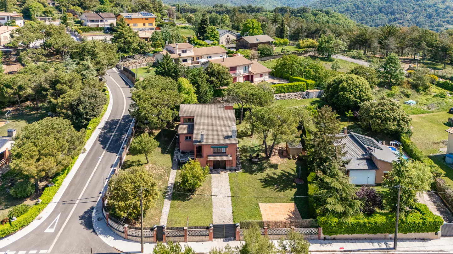 Aerial view of a two-story red brick house with a gray roof, surrounded by green trees and lawns in a hilly, wooded area.