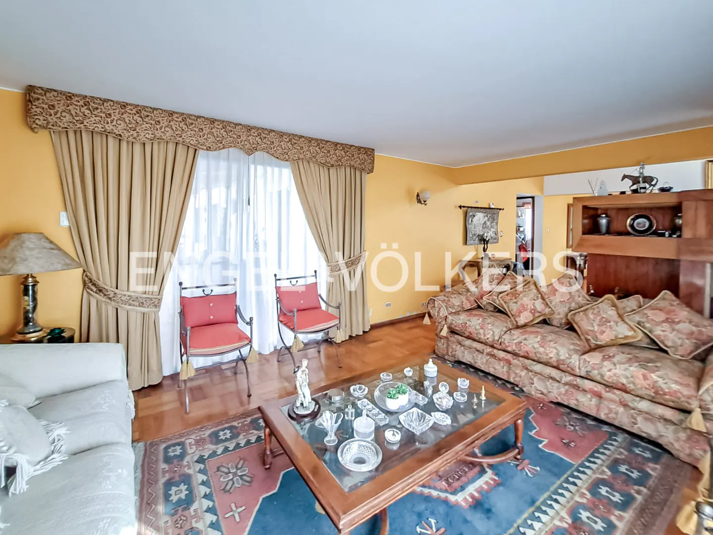 Living room with yellow walls, hardwood floors, and a blue patterned rug. Two red chairs face a window with beige curtains. A floral sofa and glass coffee table are also present.
