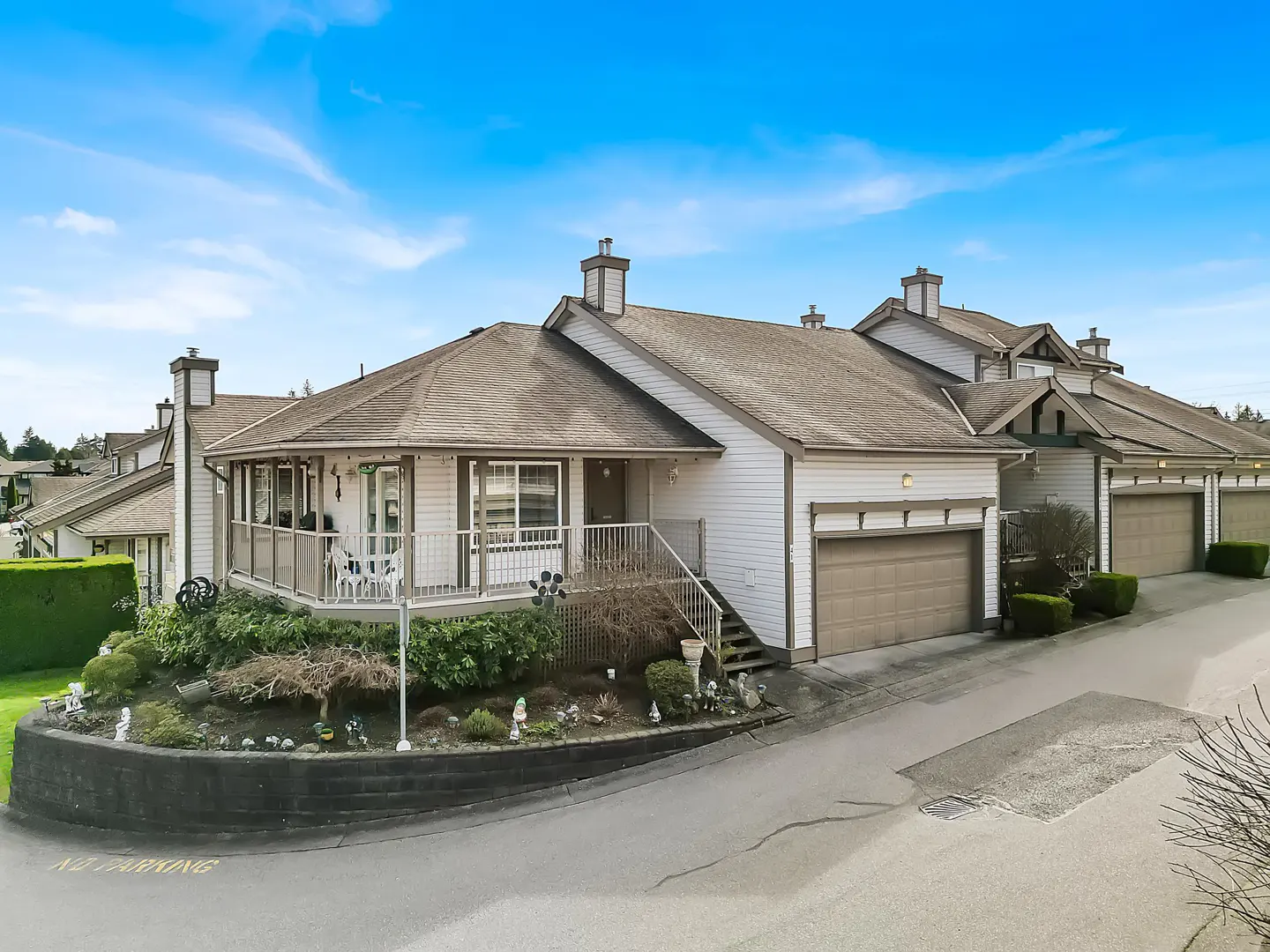 White townhouse with a wrap-around porch, brown roof, and attached garage, set against a bright blue sky.