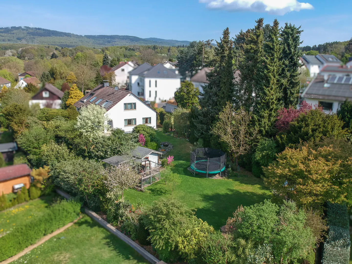 Aerial view of a white house with a brown roof, surrounded by green trees and a lawn with a trampoline.