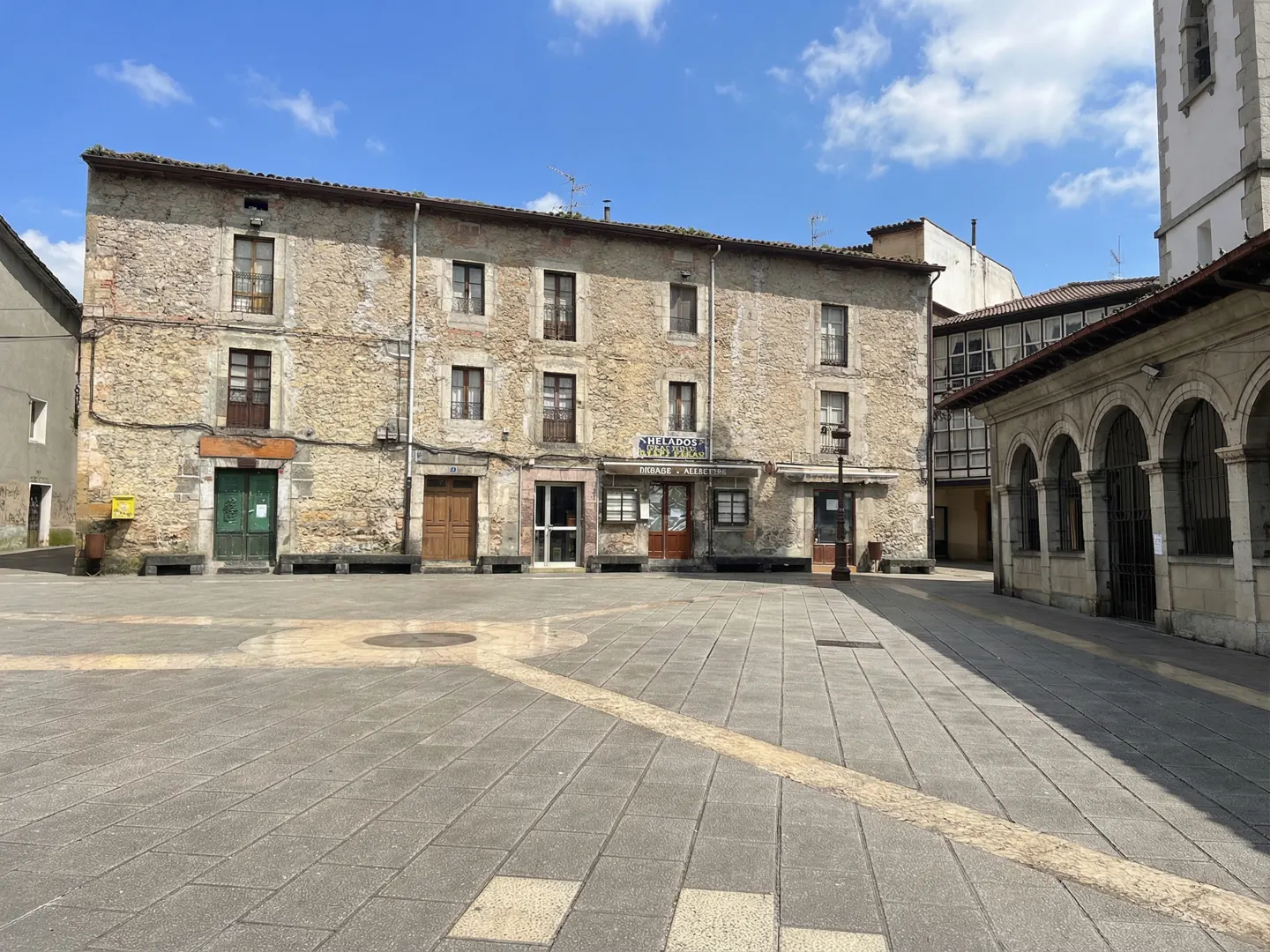 Town square with a stone building, restaurant, and arched structure under a blue sky with clouds.