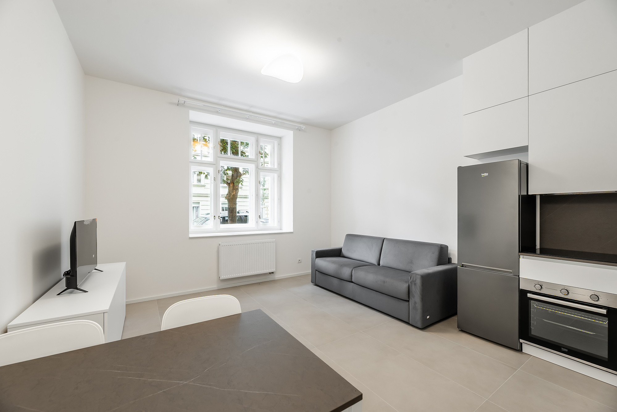 Modern studio apartment with a grey sofa, dark dining table, white cabinets, and a stainless steel refrigerator. [AI-generated text]