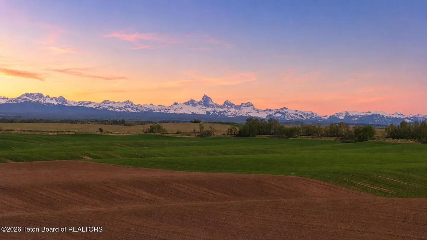 Scenic view of green and brown fields with snow-capped mountains under a colorful sunset sky.