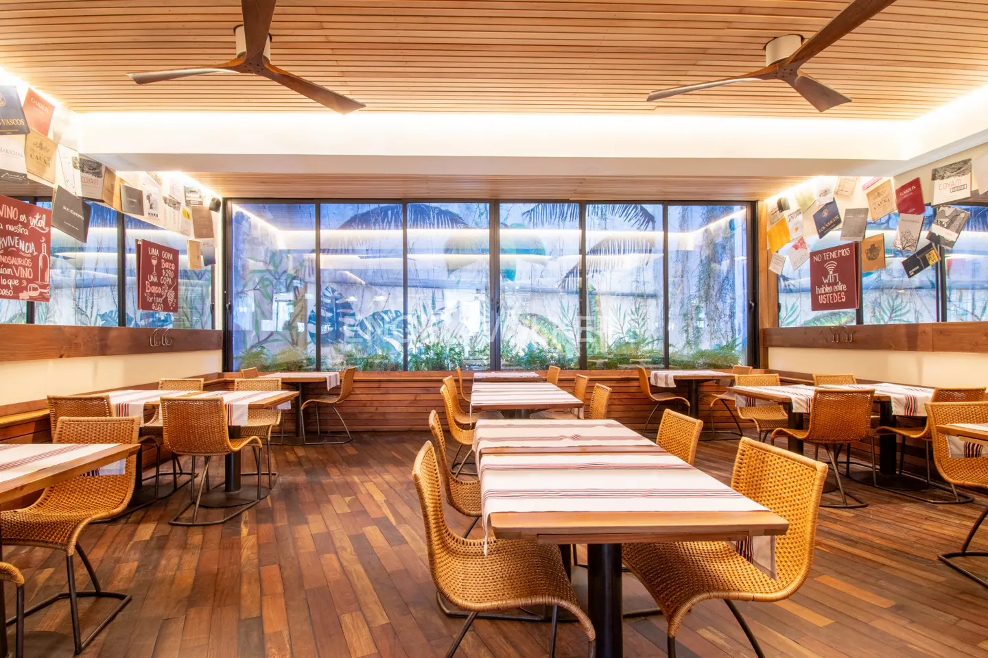 Indoor restaurant with wooden floors, tables with striped cloths, and wicker chairs. Large windows show a tropical mural. Ceiling fans and book decor add charm.