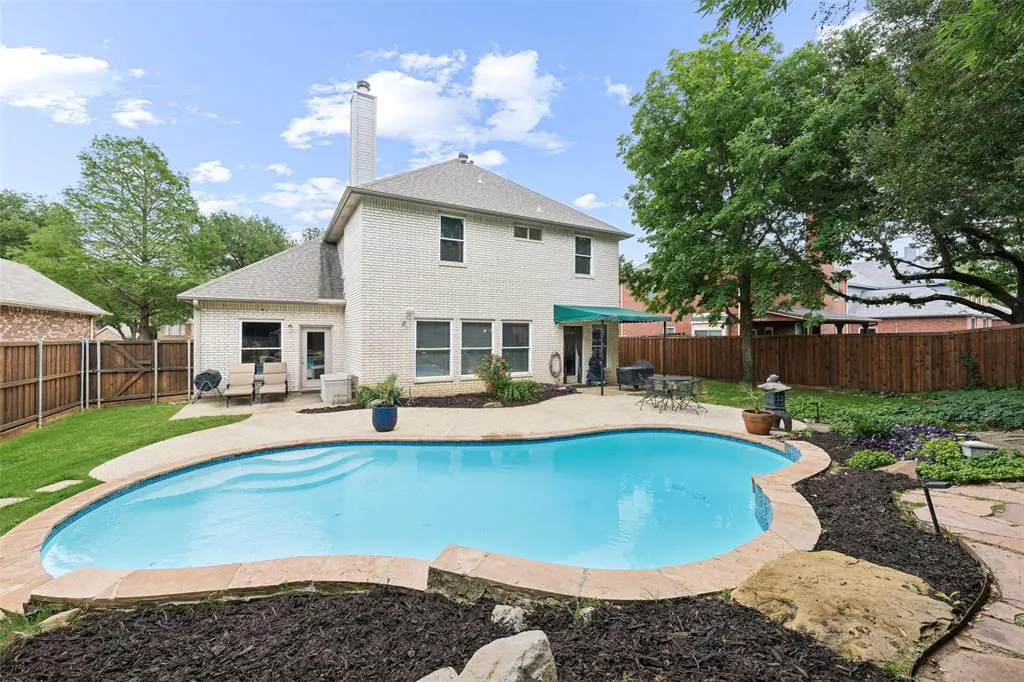 Backyard view of a two-story white brick house with a pool, patio furniture, and green trees under a blue sky.