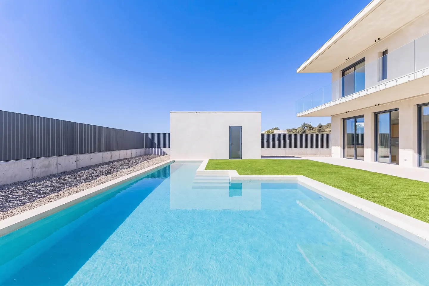 A modern house with a turquoise pool, green lawn, and white building under a clear blue sky.