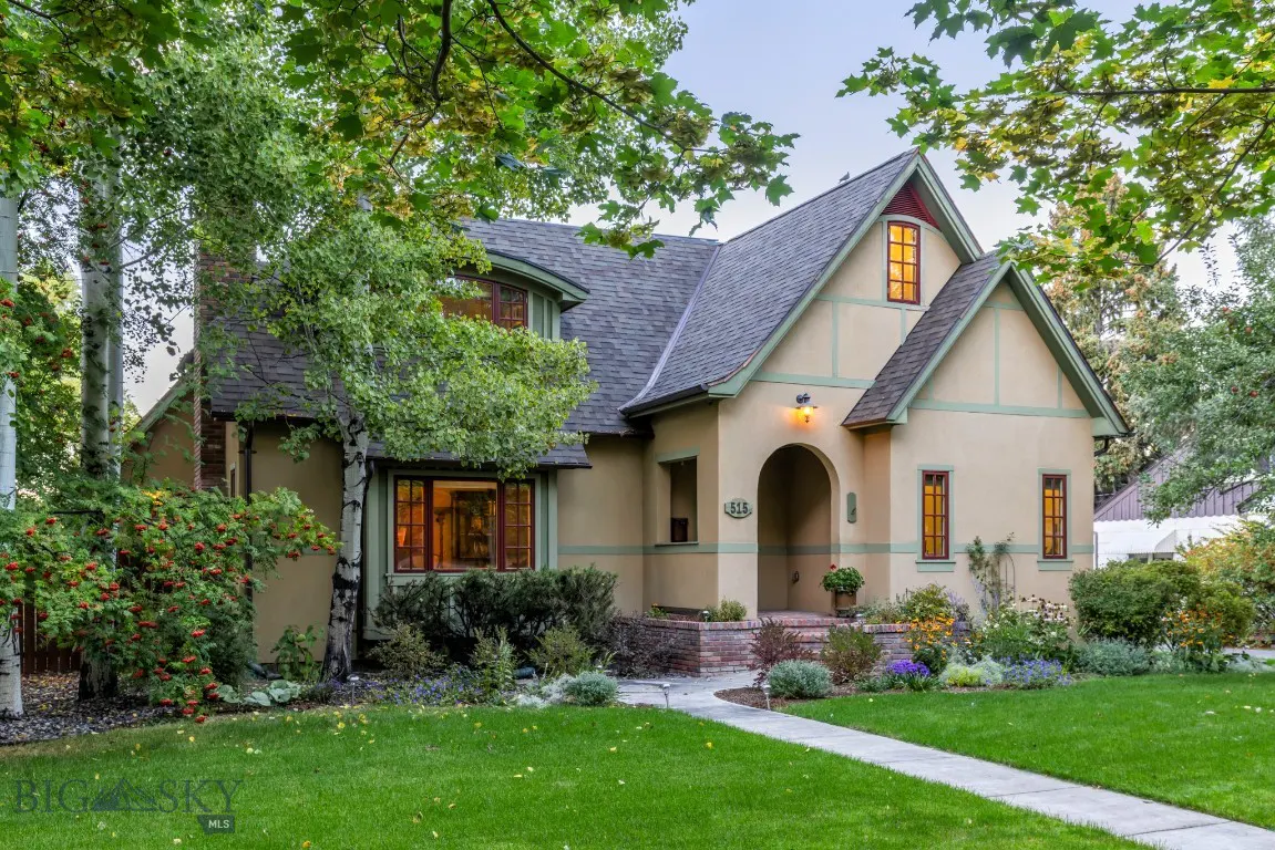 A tan Tudor-style house with green trim, a gray roof, and a brick walkway leading to the front door.
