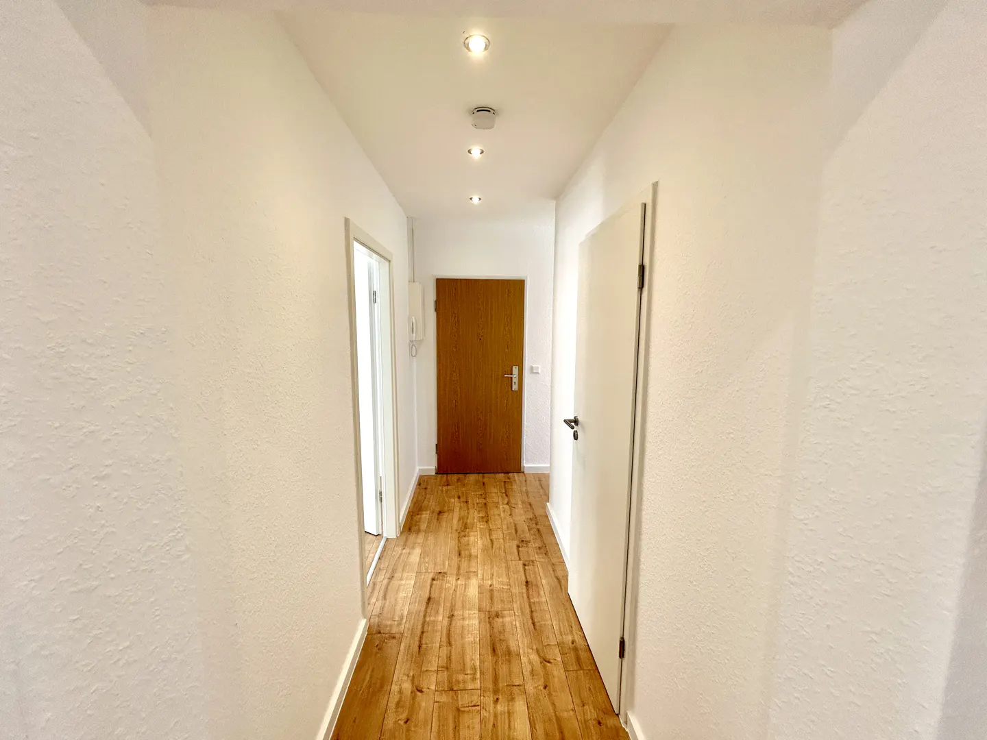 Hallway with white walls, wood floor, and recessed lighting. A brown door is at the end of the hall.