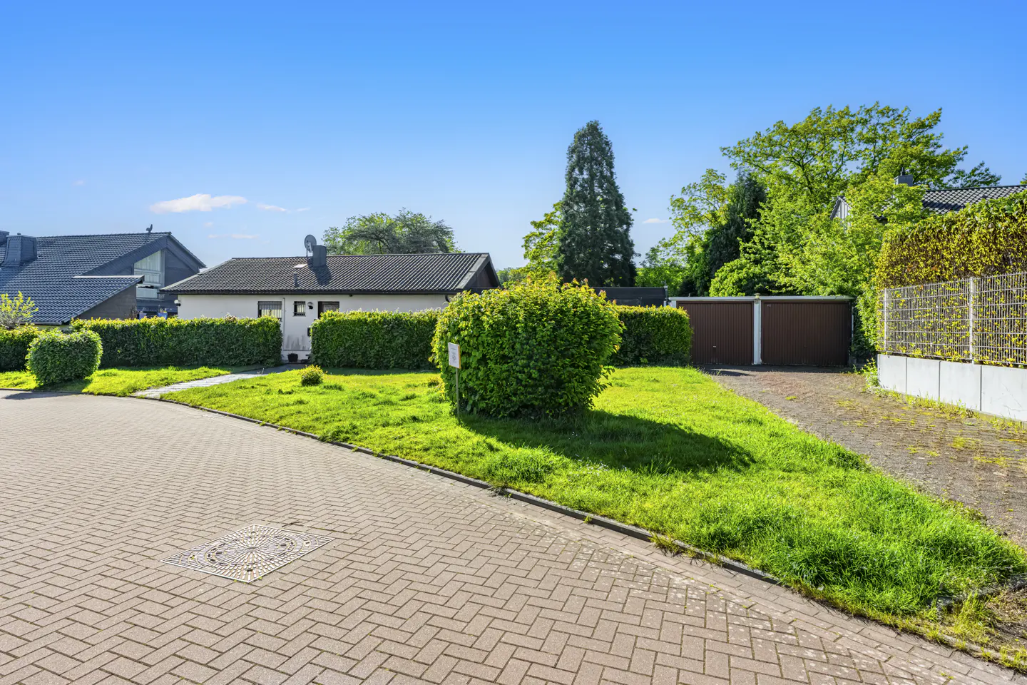 A suburban street view with brick paving, green lawns, trimmed hedges, and houses under a clear blue sky.