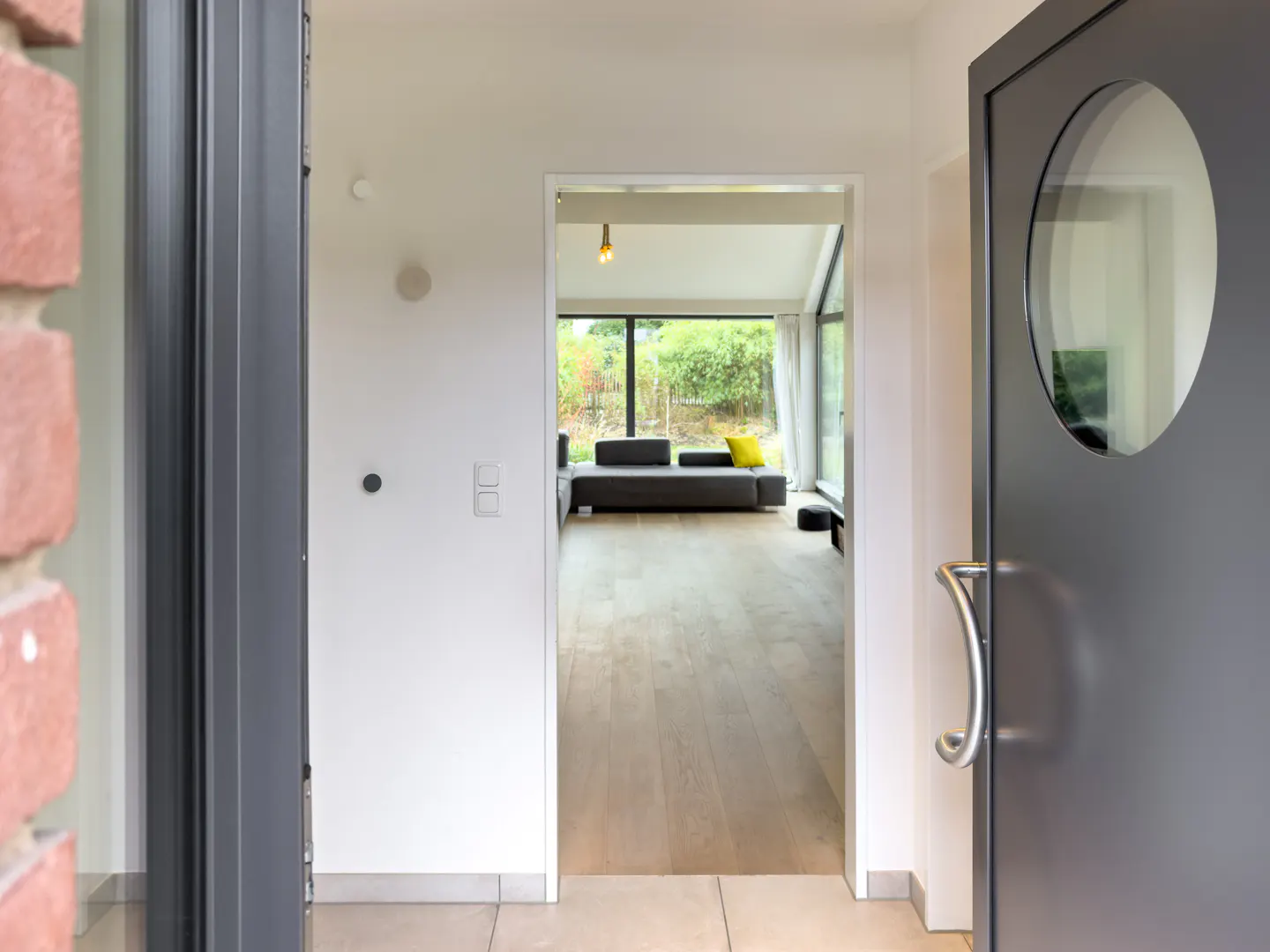 Open gray front door with round window, showing a living room with gray sofa and yellow pillow.
