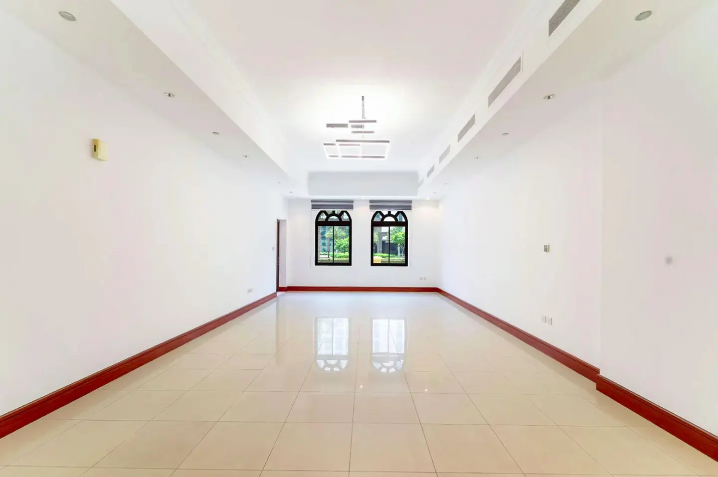 Bright, empty room with white walls, tile floor, and dark wood trim. Two arched windows show green trees outside. Modern light fixture on ceiling.