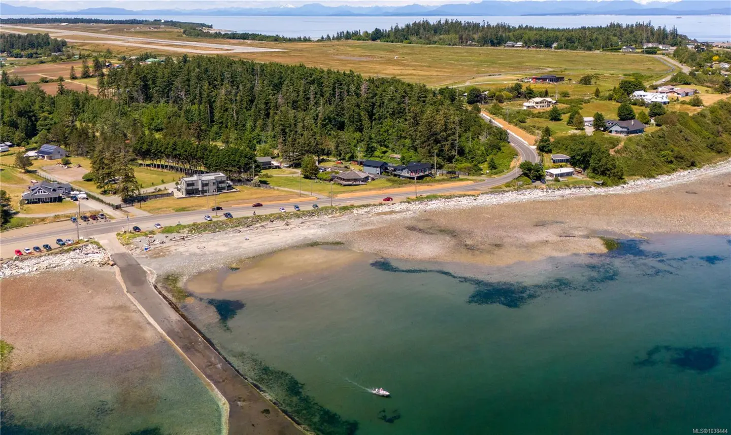 Aerial view of a coastal landscape with houses, trees, and a road along the beach. A boat is on the water.
