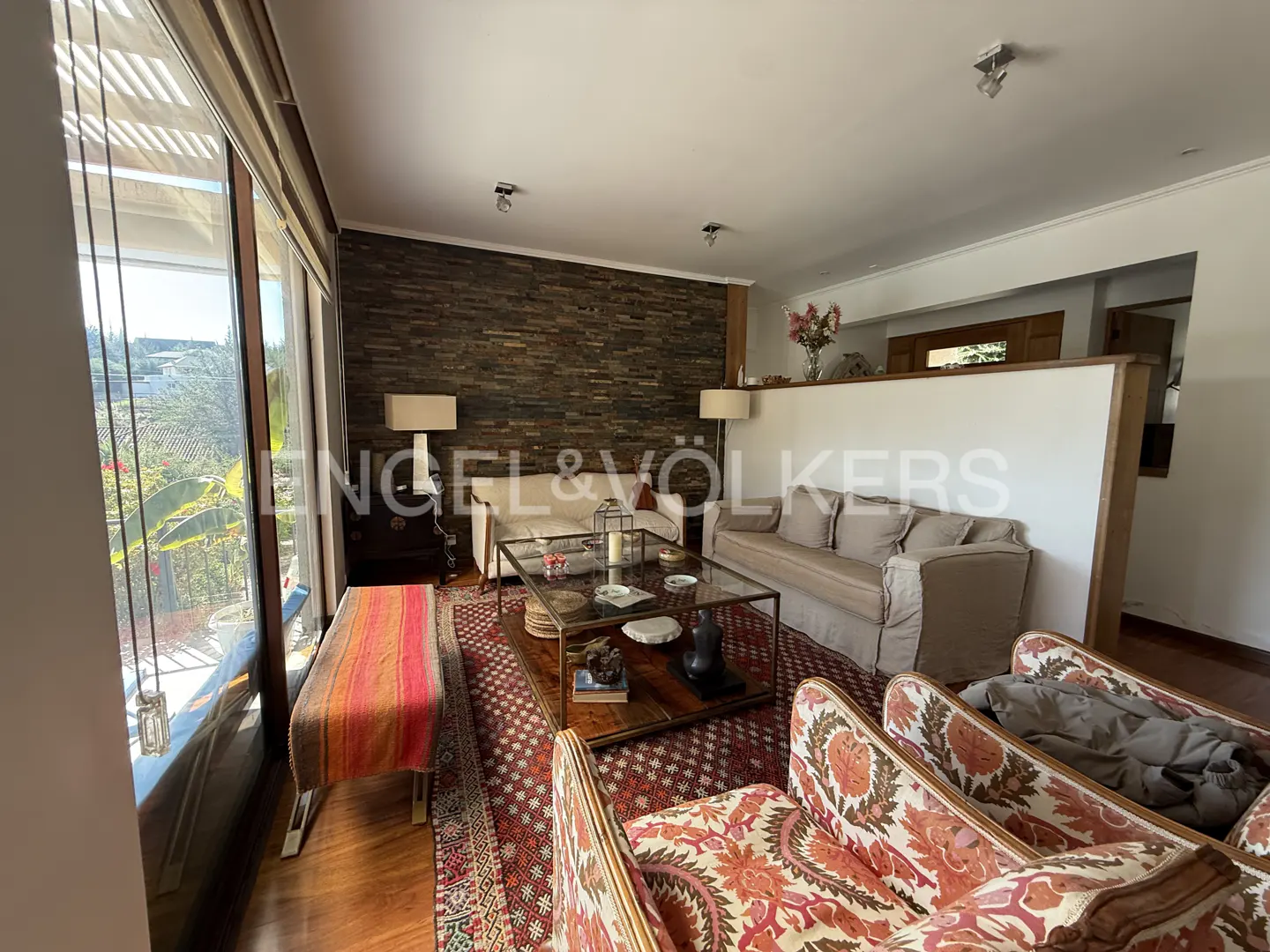 Living room with stone wall, sofas, patterned chairs, and a glass coffee table on a red rug. Large windows offer a view of greenery.